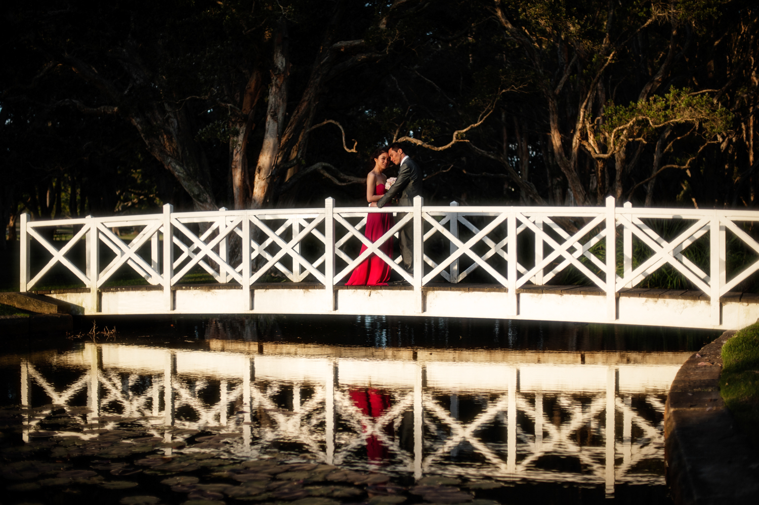 A beautiful bride and groom embracing on a bridge over a serene pond, captured by Mario Zmudzinski at Magnum Studio